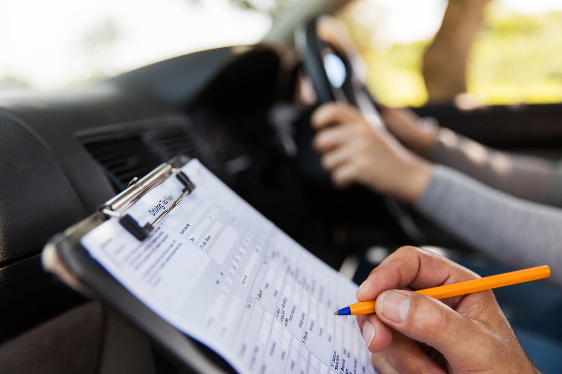 Ein Klemmbrett mit einem Formular und einer Hand, die einen Stift hält, während eine andere Person im Hintergrund ein Auto lenkt.