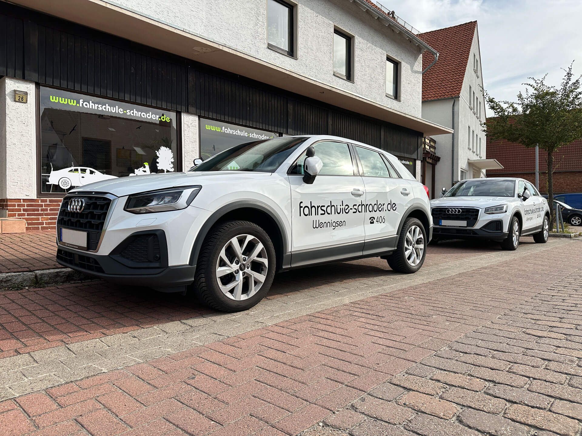 Zwei Audi-Fahrschulautos mit Markenzeichen parken vor einem Gebäude mit Fahrschulbeschriftung.
